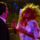 Kein Text erkennbar. Eine Person mit verwuscheltem Haar und blutbespritztem, zerrissenem Hemd steht einer anderen gegenüber.