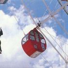Ein Mann springt aus einer roten Gondel eines großen Riesenrads in die Luft, umgeben von blauem Himmel und weißen Wolken.