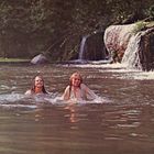 Zwei Menschen baden lächelnd in einem Fluss vor einem Wasserfall, umgeben von üppiger Vegetation.
