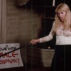 „DOWN WITH MALE SUPREMACY“ auf einem Plakat. Frau zeigt mit einem Stock auf eine pinke Statue.