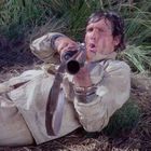 Ein Mann in heller Kleidung liegt auf dem Boden im Gras, zielt mit einem Gewehr und wirkt konzentriert.