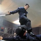 Ein Mann in einem blauen Anzug steht kampfbereit auf einem anderen, liegenden Mann in dunkler Kleidung.