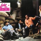 „Liebe gegen Barzahlung“ in pink; vier Männer auf Sandsäcken, rauchend; eine Frau steht; dunkler Raum.