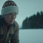 Ein älterer Mensch trägt eine Strickmütze mit roten und weißen Mustern, im Hintergrund ein verschneiter Wald.