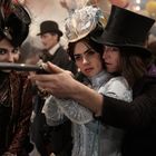 Zwei Menschen in viktorianischer Kleidung mit Hüten, einer hält eine Waffe. Eine dritte Person beobachtet grinsend.