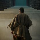 Ein Mann in einem groben Gewand steht vor einer imposanten steinernen Treppe in einem dunklen, gewölbten Raum.