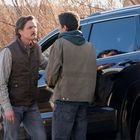 Zwei Männer stehen draußen vor einem schwarzen Auto, einer trägt eine braune Weste, der andere eine grüne Jacke.