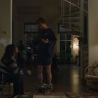 Ein Wohnzimmer mit zwei Menschen. Eine Person sitzt, die andere steht mit heruntergelassener Hose und spricht.