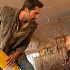 Zwei Männer in angespannter Szene: Einer steht mit gelber Flasche in Händen, der andere sitzt blutend am Boden gegen eine Wand.