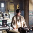 Ein Mann in historischem Gewand schreibt mit einer Feder an einem Tisch in einem Raum mit Säulen und verzierten Wänden.