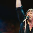 Ein Mann singt enthusiastisch in ein Mikrofon und hebt den Arm. Er trägt ein schwarzes Hemd. Bunte Lichter im Hintergrund.