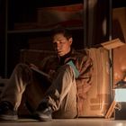 Ein Mann sitzt mit einem Buch auf dem Boden in einem schwach beleuchteten Raum, neben ihm eine Laterne.