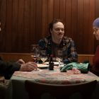 Drei Personen sitzen an einem Tisch mit Weingläsern und Tellern. Eine Frau trägt eine lila Mütze. Holzwand im Hintergrund.