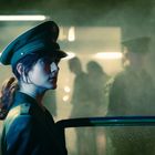 Eine Frau in Uniform mit Schirmmütze steht neben einer Autotür, im Hintergrund diffuse Gestalten und gedämpfte Beleuchtung.