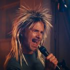 Keine Texte sichtbar. Ein Mann trägt eine wilde Perücke und singt leidenschaftlich in ein Mikrofon. Bunte Lichter im Hintergrund.