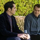 Zwei Männer sitzen nebeneinander. Einer hält ein Weinglas. Grüne Büsche im Hintergrund.