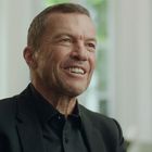 Ein Mann in einem schwarzen Anzug lächelt. Hinter ihm sind große Fenster mit grünem Laub zu sehen.