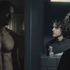 Ein Mann mit nacktem Oberkörper steht im Halbdunkel. Zwei Männer spiegeln sich in einem beleuchteten Badezimmerspiegel.