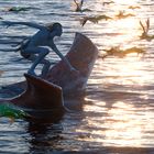 Signatur unten links. Eine blaue Figur surft auf einem großen Meereswesen, umgeben von fliegenden Kreaturen im Sonnenlicht.