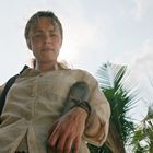 Eine Frau in heller Kleidung mit Speer vor Palmen, von unten fotografiert.