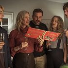 Eine Familie mit Sektgläsern singt Weihnachtslieder vor einem geschmückten Baum, ein rotes Buch mit Noten wird gehalten.