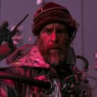 Keine sichtbaren Texte. Mann mit Bart trägt einen Helm mit Kabeln und schaut ernst. Im Hintergrund schweben Objekte.