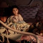 Ein kleines Mädchen sitzt im Bett, umgeben von Kissen mit Libellenmotiv und einer bunten Decke. Eine Lampe spendet warmes Licht.