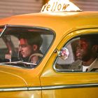 Auf einem gelben Taxi steht das Wort "Yellow", innen ein Schild "VACANT". Zwei Männer sitzen darin.