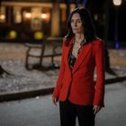 Ich kann keine genauen Details oder Personen identifizieren. Eine Frau steht draußen in der Nacht, trägt einen roten Blazer.