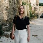 Eine Person steht vor einer alten Steinmauer in einer sonnigen Umgebung, trägt schwarze Kleidung und schaut in die Kamera.