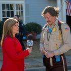 Eine Reporterin mit einem Mikrofon mit „15“ spricht mit einem Mann in Uniform vor einem Haus mit US-Flagge.