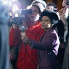 „12“ auf einem Mikrofon. Eine Reporterin mit Afrofrisur in einem lila Blazer hält das Mikrofon, umgeben von Fotografen.