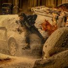 Ich kann den Text nicht identifizieren. Eine Person springt in einer verschneiten Straße neben Autos, mit Explosion und Trümmern.