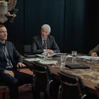 Drei Männer in Anzügen sitzen an einem Konferenztisch, mit einem US-Adler-Wappen an der Wand. Papierstapel und Gläser sind verteilt.