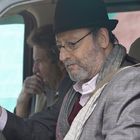 Ein Mann mit Hut und Brille sitzt am Steuer eines Fahrzeugs. Ein weiterer Mann sitzt auf dem Beifahrersitz.