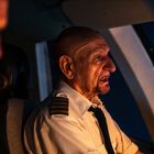 Ein älterer männlicher Pilot in Uniform sitzt im Cockpit eines Flugzeugs, abends beleuchtet.