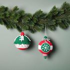 Zwei gehäkelte Weihnachtskugeln in Rot, Grün und Weiß hängen an einem Tannenzweig vor einem grauen Hintergrund.