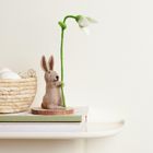 Ein Stoffhase hält eine Filz-Schneeglöckchenblume. Links steht ein Korb mit Eiern auf einem Buchstapel.
