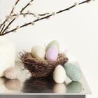 Ein weißer Krug mit Weidenzweigen, ein Nest mit pastellfarbenen Eiern aus Filz auf einem Tisch.
