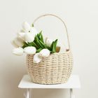 Ein geflochtener Korb mit weißen Tulpen steht auf einem weißen Tisch vor einer hellen Wand.