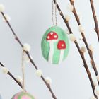 Filz-Osterei mit zwei roten Pilzen auf grünem Hintergrund, hängend an einem Zweig mit flauschigen Knospen.