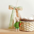 Stoffhase in grünem Outfit mit Karotte, daneben ein Korb mit Eiern und ein Farbband, das zu einer Schleife gebunden ist.