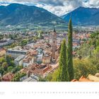 3 • 2027. Eine malerische Stadtansicht mit Kirchturm, umgeben von Bergen und grüner Landschaft.