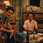 Texte: "PILSENER", "PREMIUM QUALITY". Eine Gruppe von sieben Menschen sitzt entspannt im Wohnzimmer, manche trinken Bier.