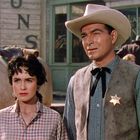 Zwei Menschen, eine Frau in kariertem Hemd und ein Mann mit Sheriff-Stern und Cowboyhut, stehen in einer Westernstadt.