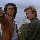 Zwei Männer stehen im Freien, einer zielt mit einer Pistole nach vorne. Sie wirken konzentriert.