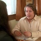 Eine Frau in beiger Bluse sitzt in einem Café und spricht mit jemandem, der im Vordergrund sitzt.