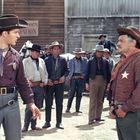 Im Hintergrund: "COACH STATION". Vorne zwei Männer in Westernkleidung und Cowboyhüten, einer mit Sheriff-Stern, umgeben von Leuten.