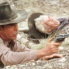 Zwei Männer in Cowboy-Outfits, einer hält einen Revolver. Sie sind in einer trockenen Landschaft liegend.