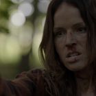 Eine Frau mit entschlossener Miene hält eine Pistole in einem Wald. Sie ist verletzt und trägt ein kariertes Hemd.